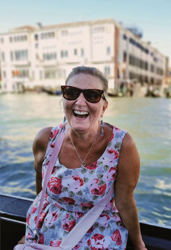 Smiling woman in sunglasses and floral dress on a boat, with a blurred waterside building in the background.