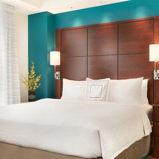 Modern hotel room with a king-sized bed, white bedding, teal accent wall, wooden headboard, and side tables with lamps; window with sheer curtains.