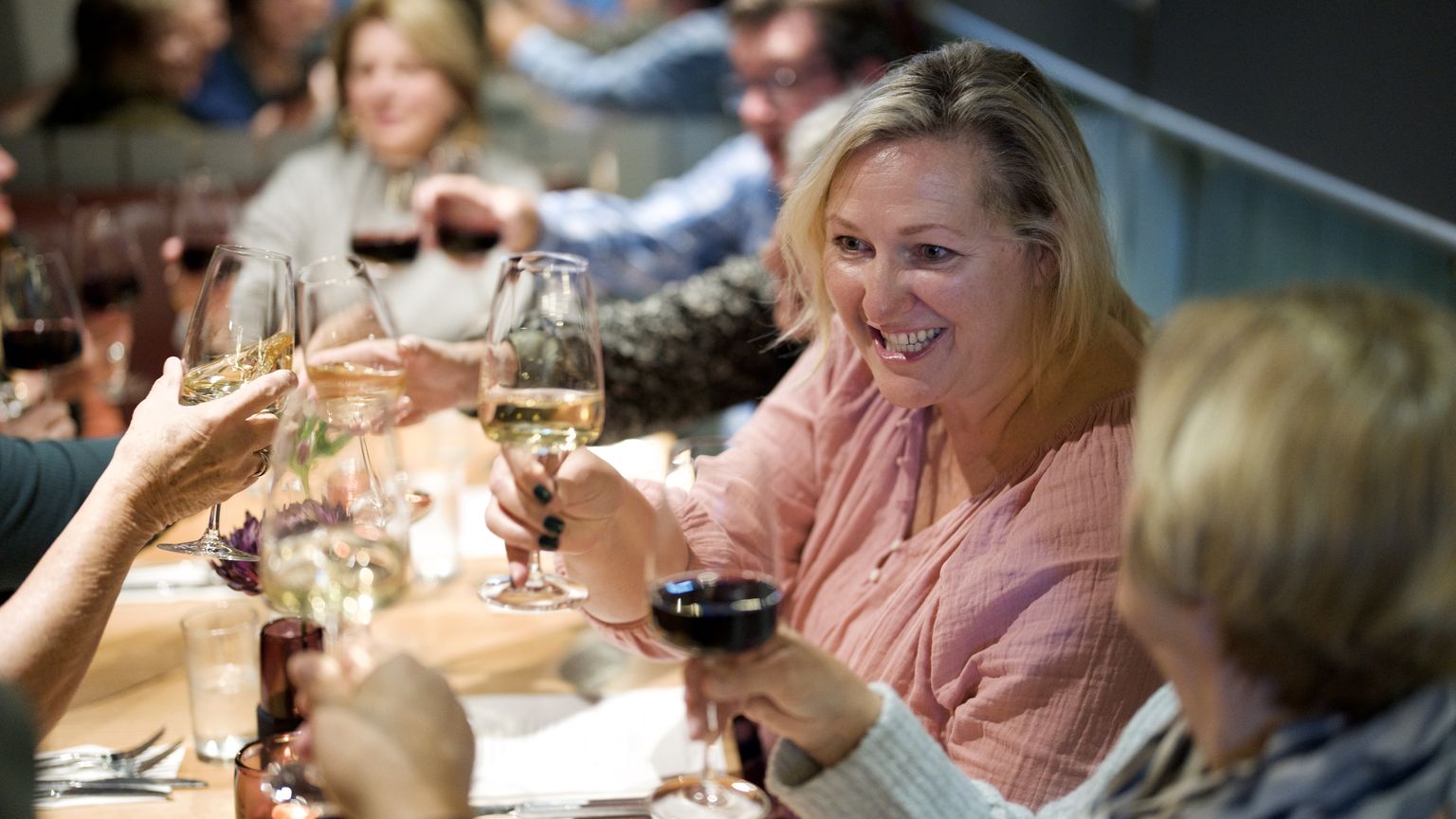 People enjoying dinner at a restaurant, raising glasses in a toast, smiling and seated around a wooden table with drinks and light food.