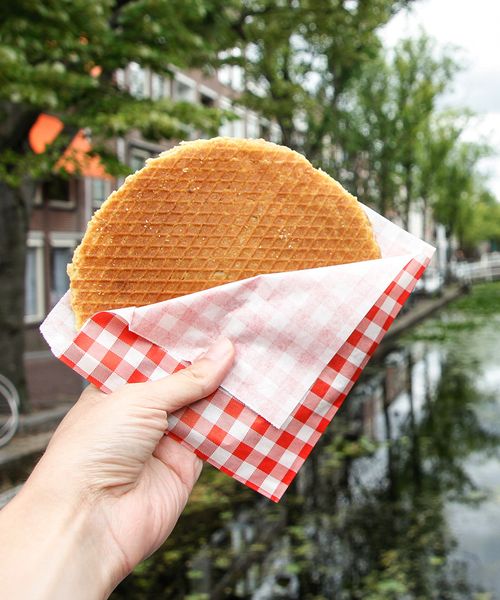 Hand holding a stroopwafel in a red-checkered napkin, with a canal and trees in the background.