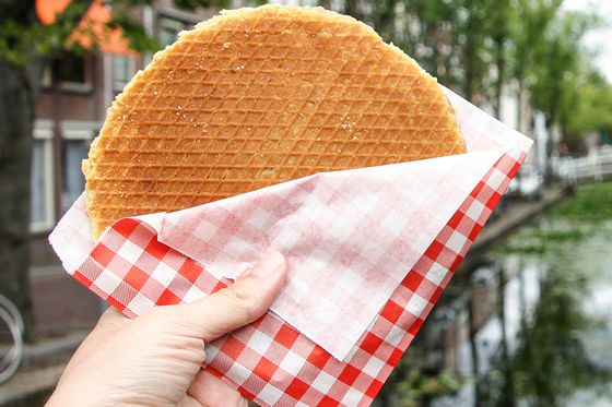 Hand holding a stroopwafel in a red-checkered napkin, with a canal and trees in the background.
