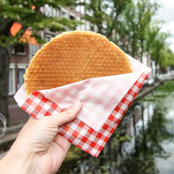 Hand holding a stroopwafel in a red-checkered napkin, with a canal and trees in the background.