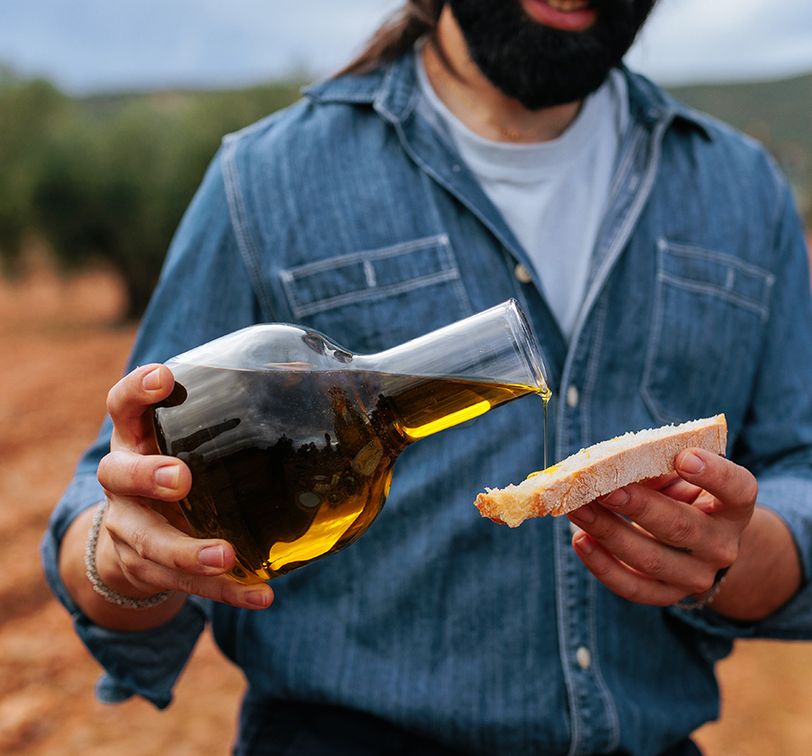 Man pouring olive oil from a carafe onto a slice of bread outdoors, wearing a denim shirt, with blurry trees in the background.