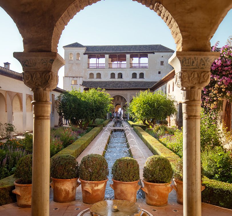 A beautiful stone courtyard with a central water feature, framed by ornate arches and columns, leads to a historic building surrounded by lush greenery.