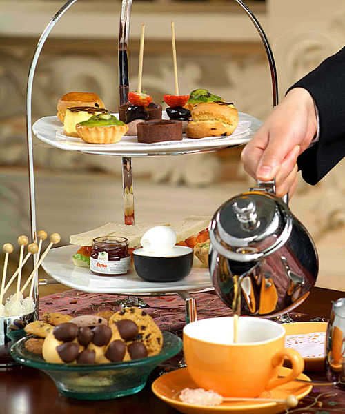 A two-tiered tray with assorted pastries and savories. A person pours tea into a cup beside cookies and condiments.