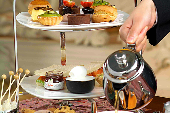 A two-tiered tray with assorted pastries and savories. A person pours tea into a cup beside cookies and condiments.