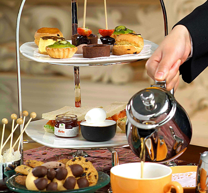 A two-tiered tray with assorted pastries and savories. A person pours tea into a cup beside cookies and condiments.