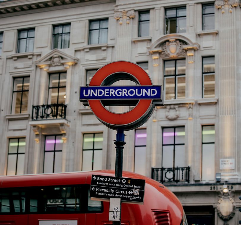 Red London Underground sign with a double-decker bus and ornate stone building in the background.