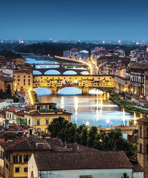 Aerial view of Florence at dusk, featuring the illuminated Ponte Vecchio and the Arno River lined with historic buildings.