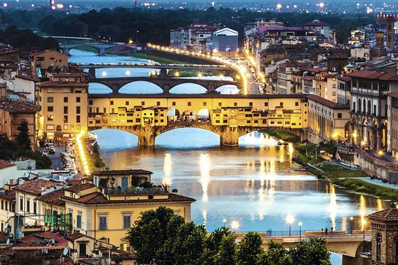 Aerial view of Florence at dusk, featuring the illuminated Ponte Vecchio and the Arno River lined with historic buildings.