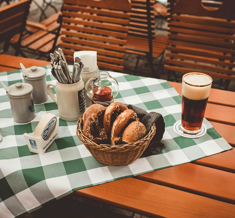 Basket of pretzels, a dark beer, condiments, and utensils on a green checkered tablecloth at an outdoor café with wooden chairs.
