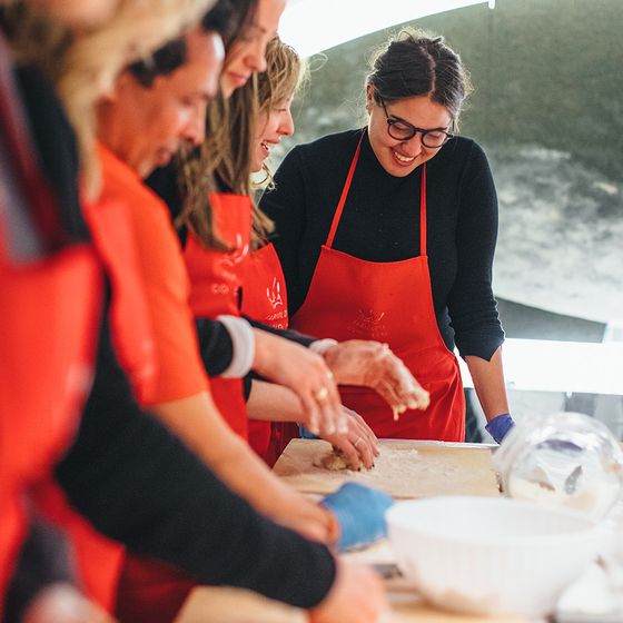 A group of people wearing red aprons are gathered around a table, smiling and making dough together.