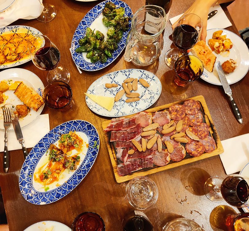 A table filled with various tapas dishes, including meats, cheese, brussels sprouts, and wine glasses, surrounded by people enjoying the meal.