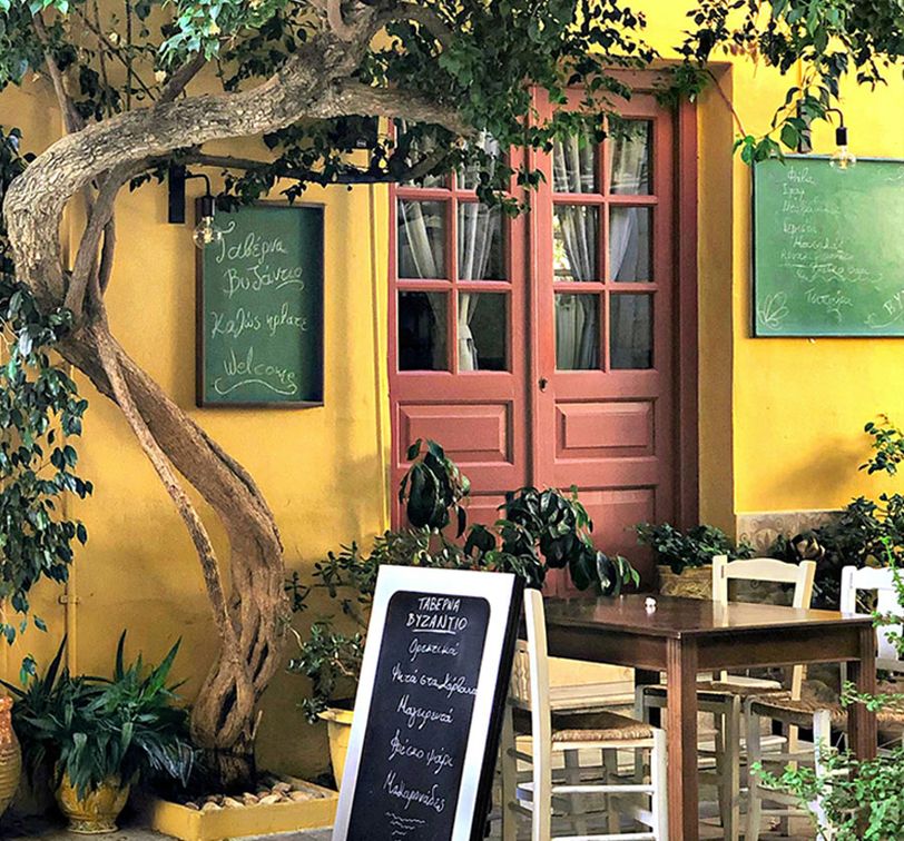 Charming outdoor café with a yellow wall, pink door, chalkboard menus, wooden tables, and chairs under a leafy tree.