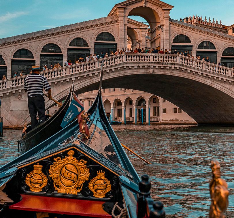 A gondola approaches the ornate Rialto Bridge over a canal in Venice, Italy, with a blue sky in the background.