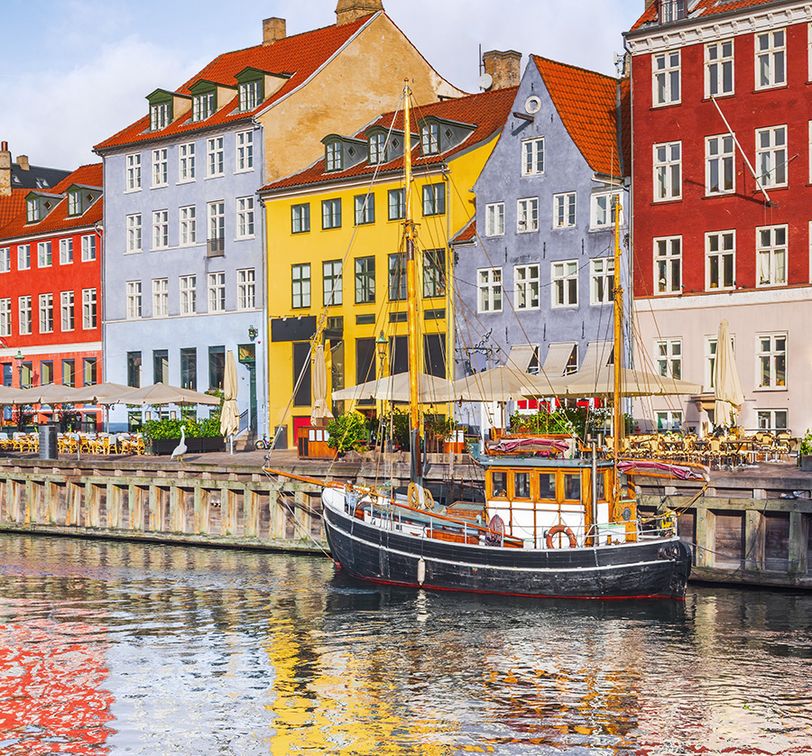 Colorful historic buildings and a moored boat reflect on the water in Nyhavn, Copenhagen, under a blue sky.