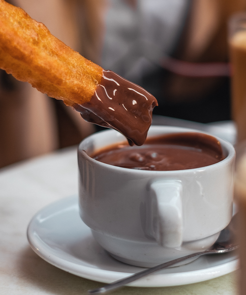 A churro dipped in a cup of thick hot chocolate on a white table with a spoon beside the cup.