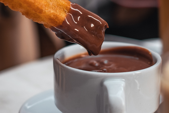 A churro dipped in a cup of thick hot chocolate on a white table with a spoon beside the cup.