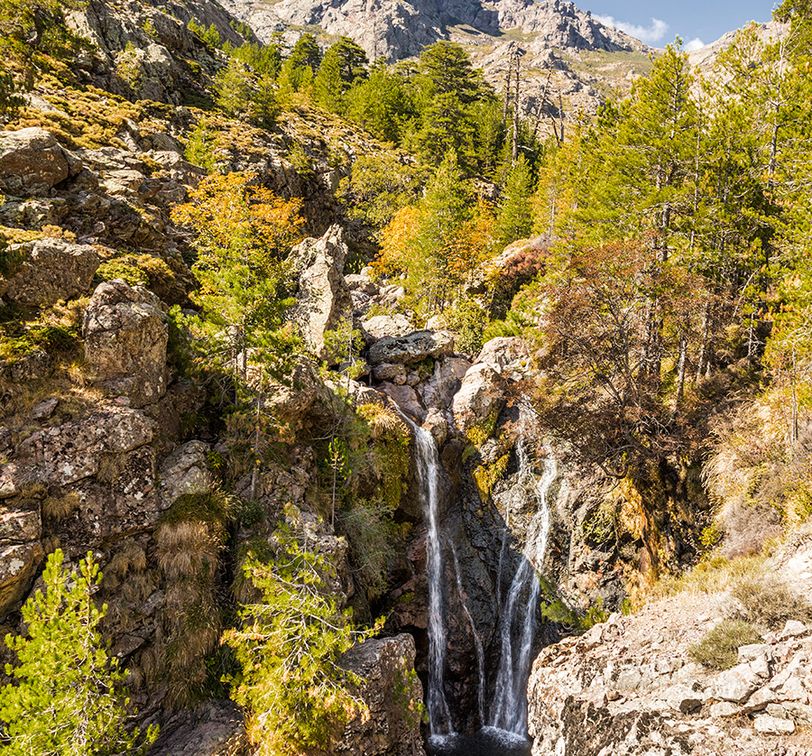 Majestic mountain landscape with a cascading waterfall surrounded by lush green trees and rocky terrain under a clear blue sky.