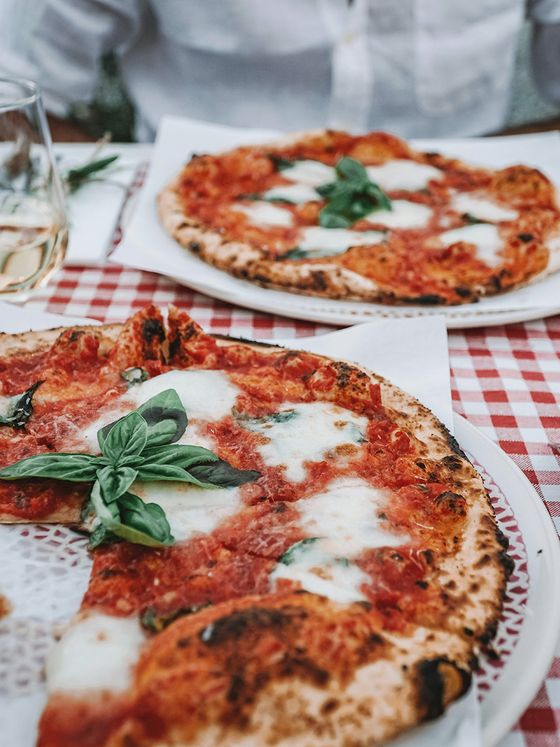 Two margherita pizzas with fresh basil and melted mozzarella on a red checkered tablecloth, accompanied by a glass of white wine.