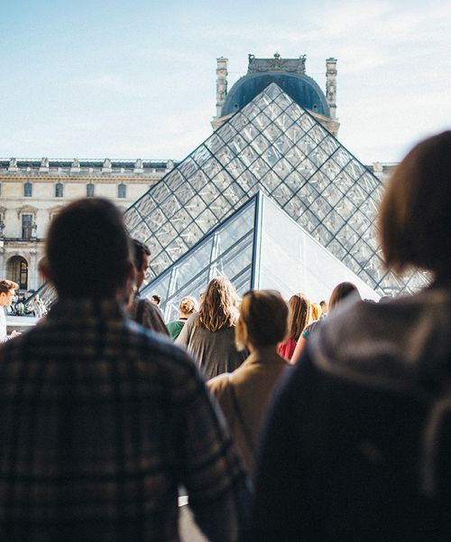 People walking towards the glass pyramid entrance of the Louvre Museum in Paris on a clear day.