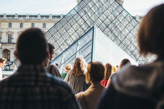 People walking towards the glass pyramid entrance of the Louvre Museum in Paris on a clear day.