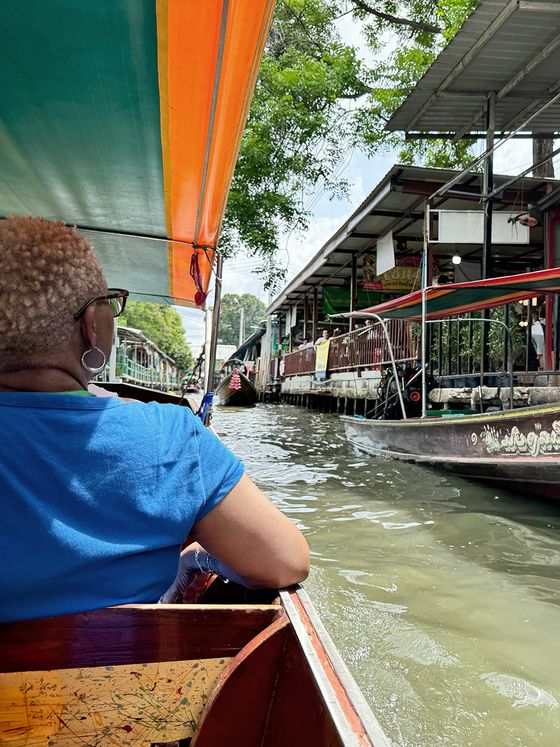 Person in a blue shirt sits in a boat navigating a canal with nearby market stalls, under a colorful canopy, on a sunny day.