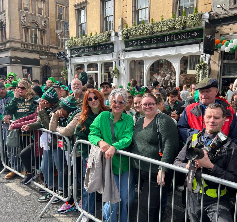 Crowd dressed in green gathers behind a barrier, celebrating in front of a shop decorated for an Irish-themed event.