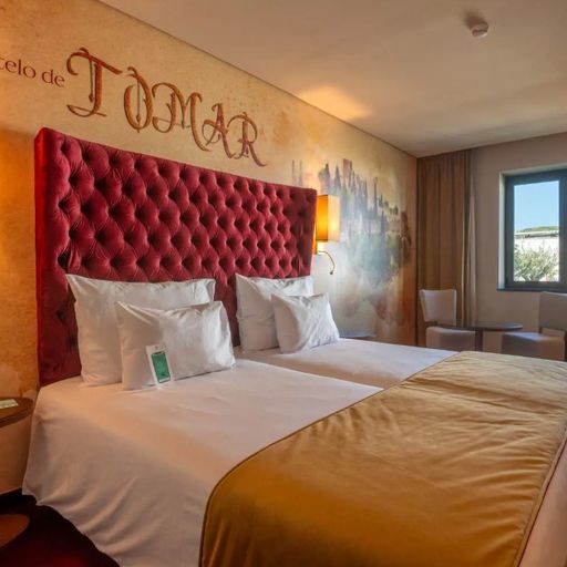 Hotel room with two beds, red tufted headboard, beige carpet, wall art featuring "Castelo de Tomar," two chairs by a window, and warm lighting.
