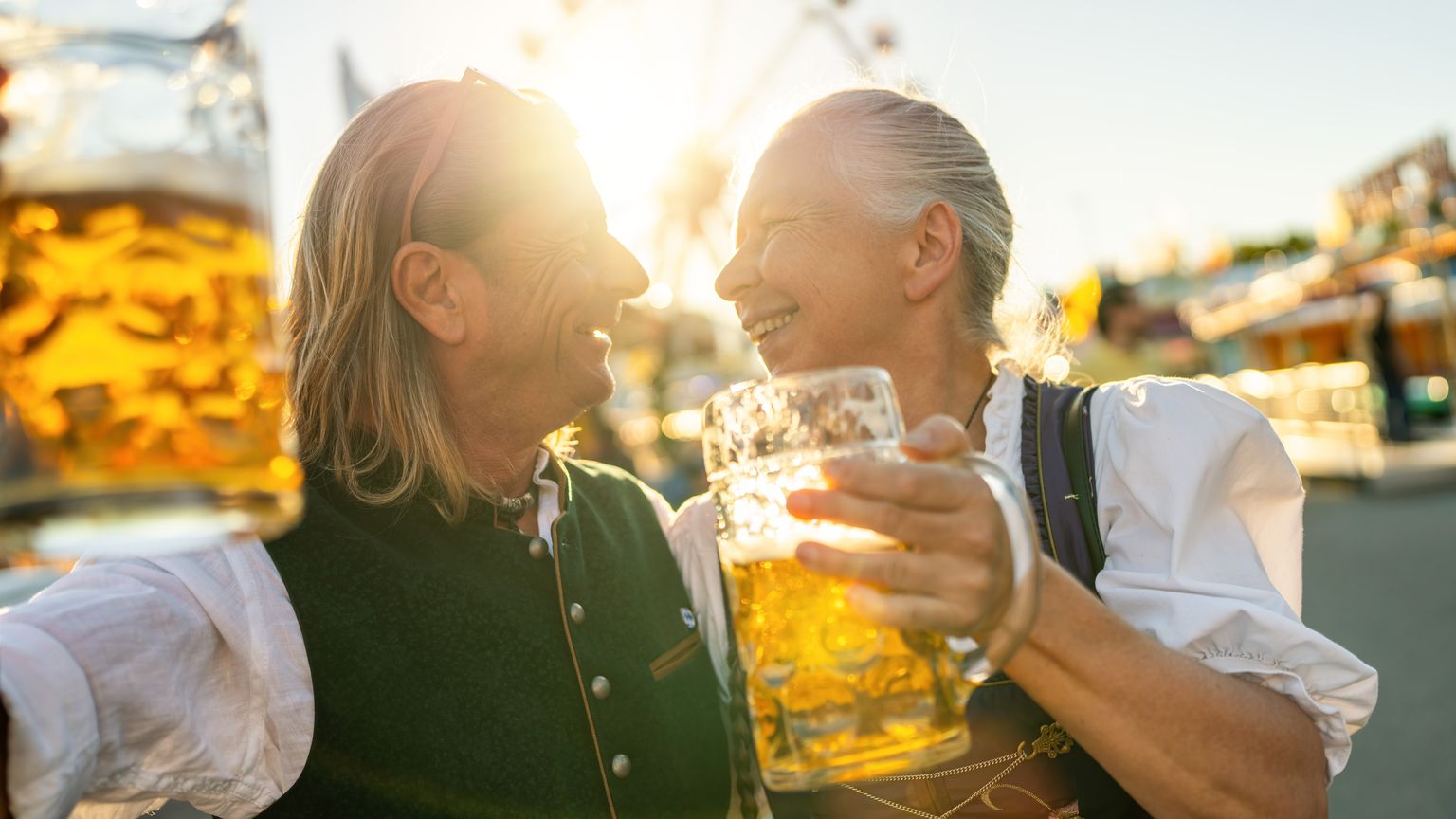 Two people in traditional attire sharing a joyful moment, holding large beer mugs with a ferris wheel in the background and sunlight filtering through.