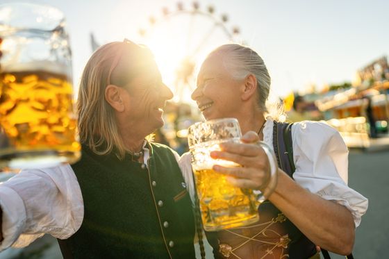 Two people in traditional attire sharing a joyful moment, holding large beer mugs with a ferris wheel in the background and sunlight filtering through.
