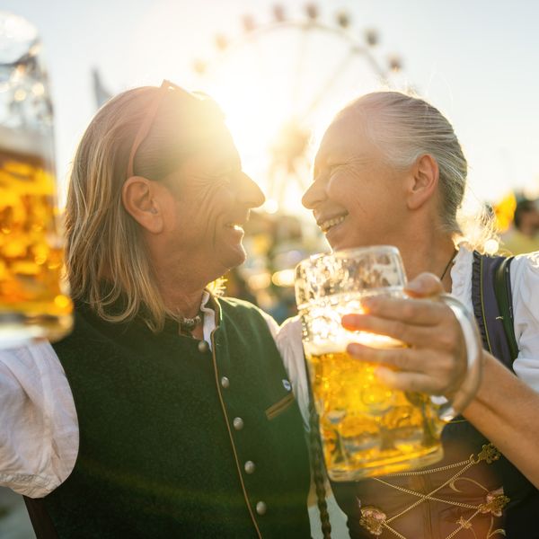 Two people in traditional attire sharing a joyful moment, holding large beer mugs with a ferris wheel in the background and sunlight filtering through.