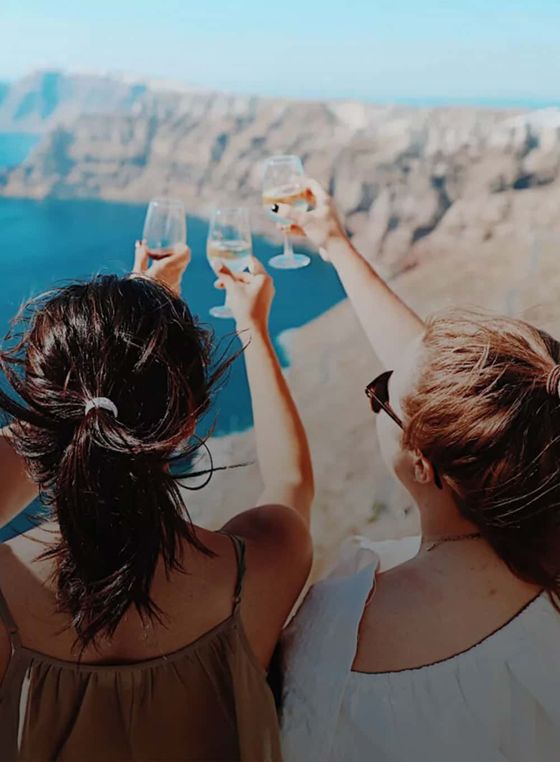 Two women raising wine glasses towards a scenic ocean and cliff view, celebrating a moment under a clear blue sky.