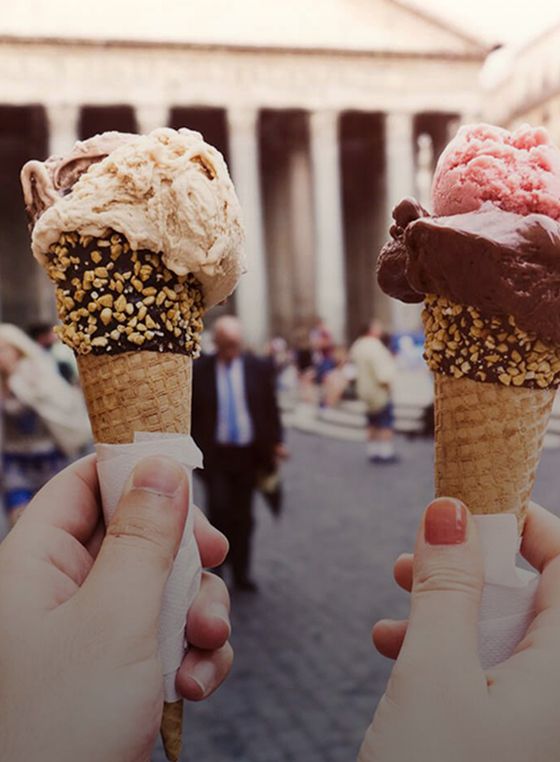 Two hands hold ice cream cones with nuts and various flavors. A blurred historical building and people are in the background.