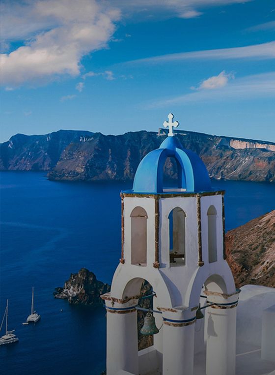 A white Greek church with a blue dome overlooks the Aegean Sea, with sailboats and a mountainous island in the background under a clear sky.