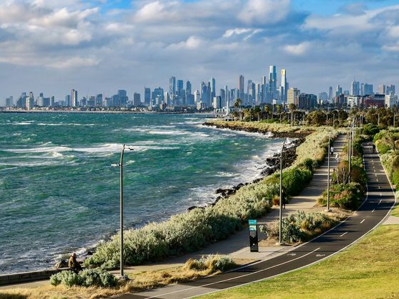 Coastal path alongside a vibrant sea, leading to a city's skyline under a partly cloudy sky.