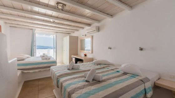 Bright, minimalist bedroom with twin beds, wooden ceiling, and open door showcasing a sea view. Towels are neatly placed on the beds.