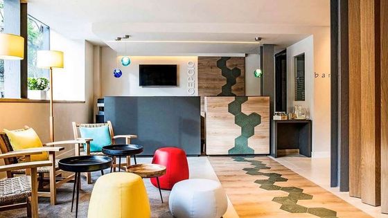Modern hotel lobby with wooden furniture, colorful stools, hexagonal-patterned floor, and large windows allowing natural light.