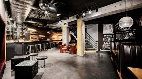 Stylish bar interior with a long counter, high stools, cushioned chairs, wall art with phrases, and modern lighting in a spacious setting.