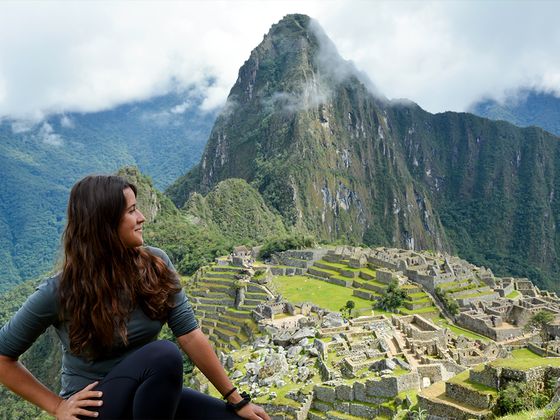 Woman sitting, smiling at the view of Machu Picchu ruins with lush green terraces and a mist-covered mountain in the background.