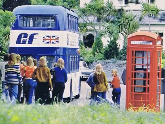 People boarding a blue double-decker bus with a British flag, near a classic red telephone box, surrounded by greenery.