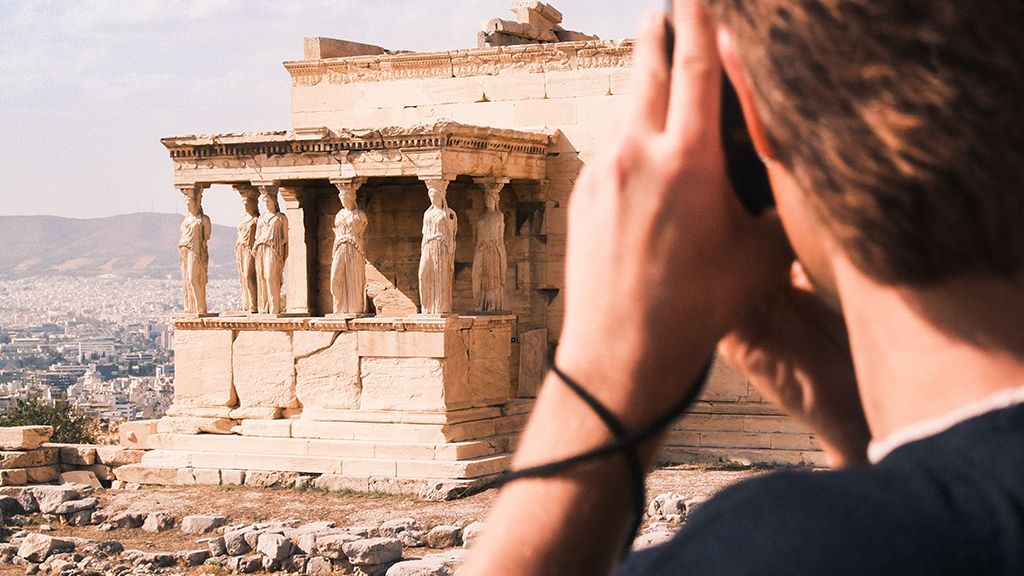 A person takes a photo of the Erechtheion with its iconic Caryatids on the Acropolis, Athens, under a partly cloudy sky.