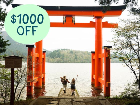 Two people walk towards a large orange torii gate by a lake, with a green circle showing "$1000 OFF" in the top left corner.