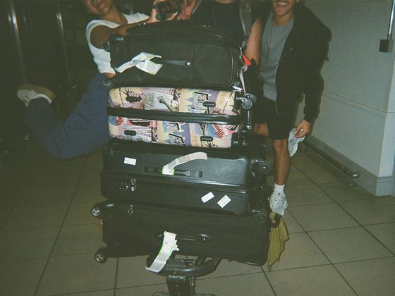 A stack of suitcases on a luggage cart, with people playfully holding onto it, in a tiled-floor setting.