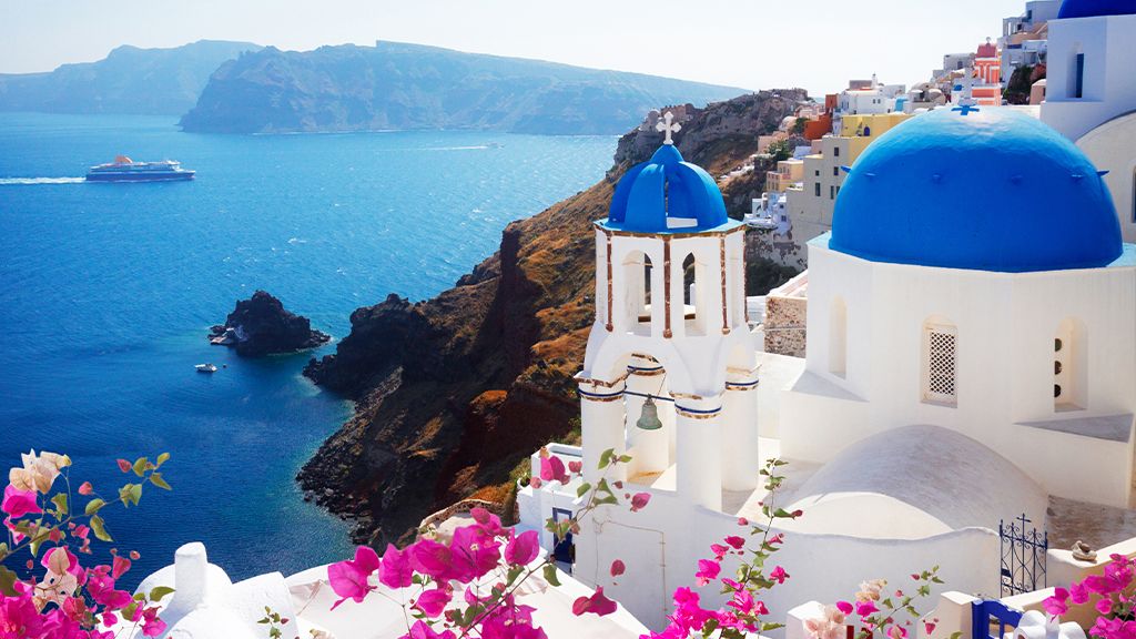 White buildings with blue domes overlooking the sea in Santorini, Greece, with pink flowers in the foreground and a ferry on the water.