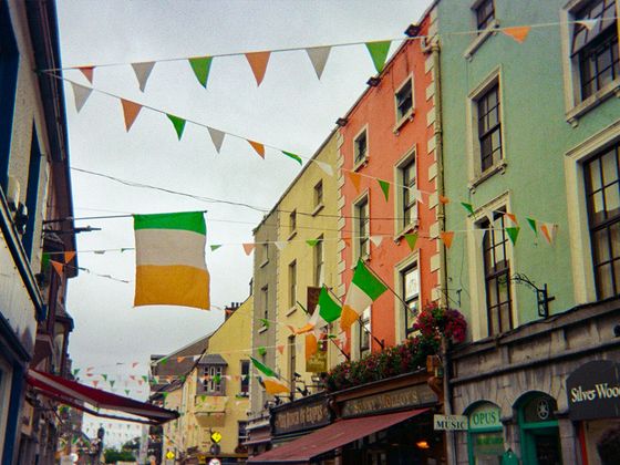 A street scene with colorful buildings adorned with Irish flags and festive buntings, creating a lively atmosphere.