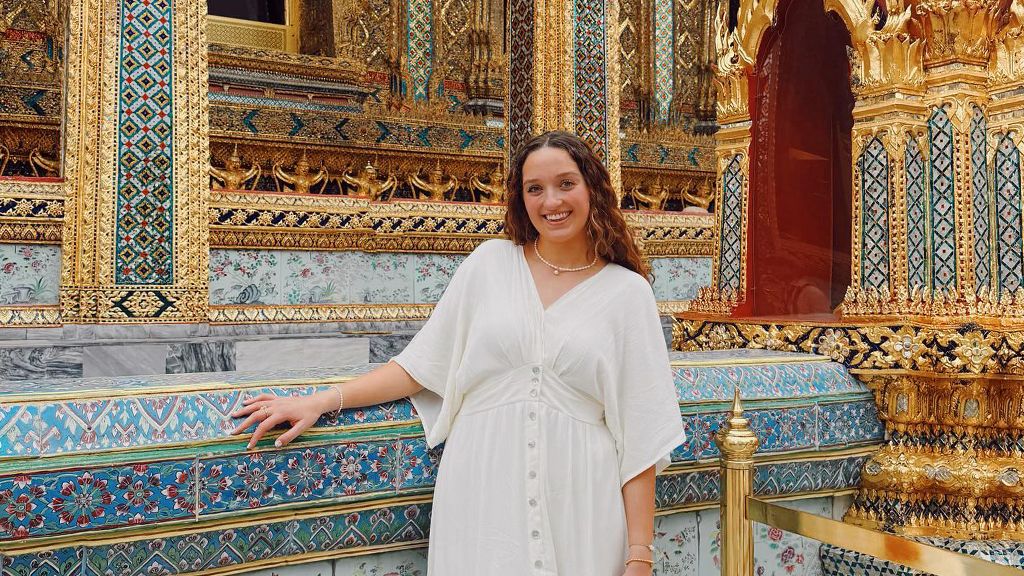 A woman in a white dress smiles while standing by ornate gold and mosaic temple decorations.