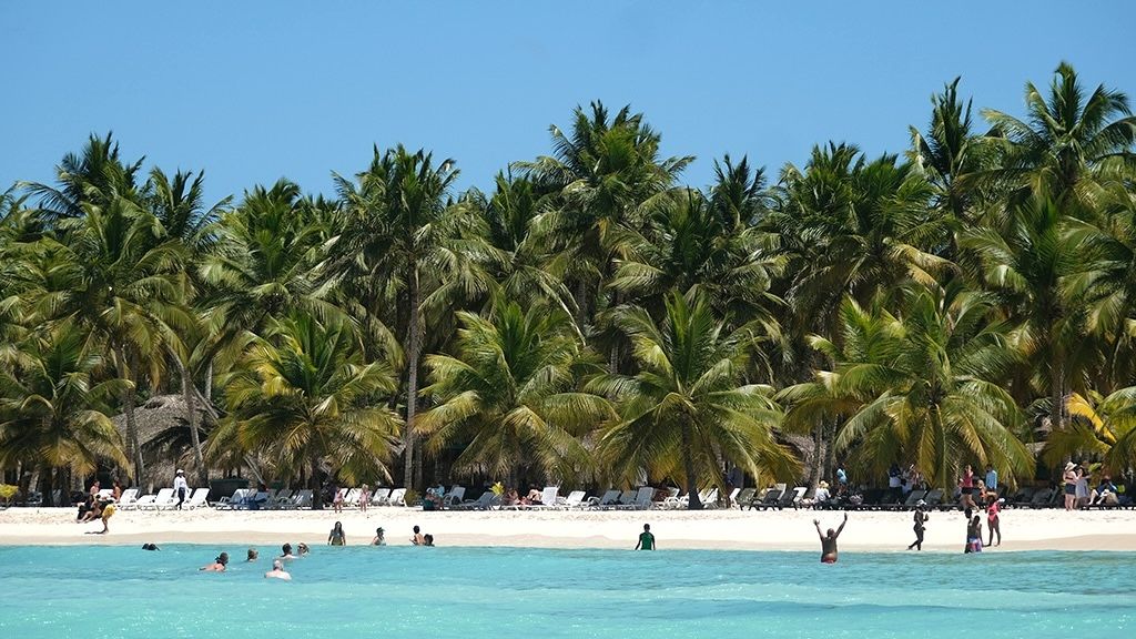 Tropical beach scene with palm trees, clear turquoise water, and people swimming and lounging on the sand under a blue sky.