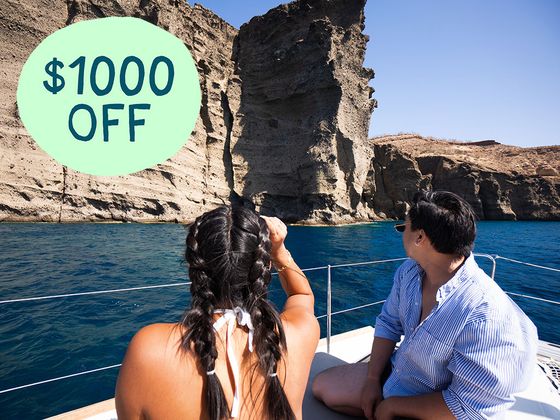 Two people on a boat observe rocky cliffs by the sea under a clear sky. A green circle in the upper left corner reads "$1000 OFF."