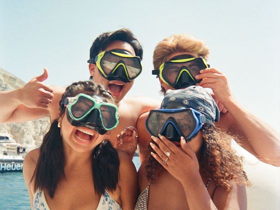 Four people wearing snorkeling masks smile and pose for a photo by the sea, with one giving a thumbs-up gesture.
